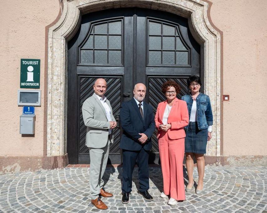 Four individuals stand in front of a building, two men and two women, dressed formally. Behind them is a large door with a 'Tourist Information' sign.