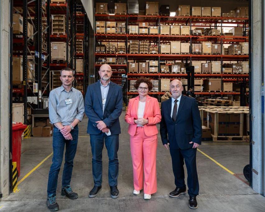 Four professionals stand in a warehouse, facing the camera. A woman in pink stands between two men in suits. Behind them, shelves filled with boxes are visible.
