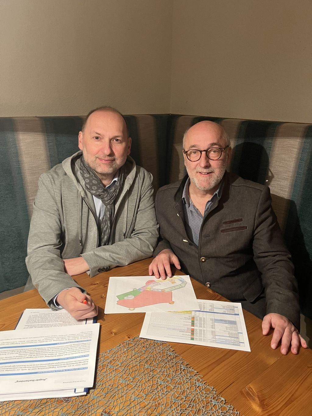 Two men sit at a wooden table in a booth. They smile for a picture. Papers and a drawing are on the table.
