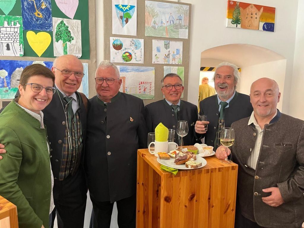 Sechs Männer in Anzügen stehen nebeneinander in einem Raum mit einem hölzernen Tresen und Weinläsern. Hinter ihnen befinden sich Gemälde an der Wand.