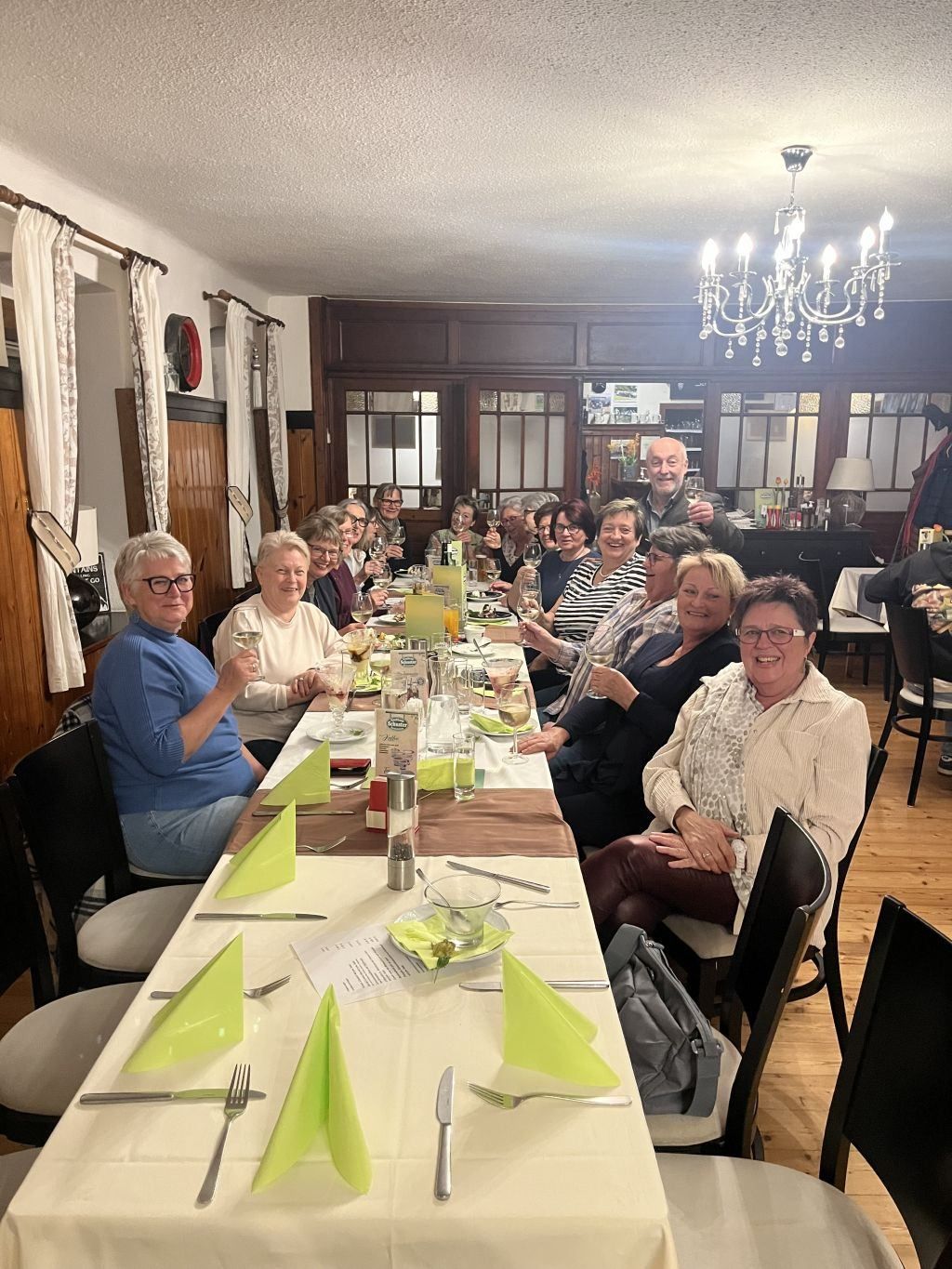 Eine Gruppe Erwachsener, einige mit Brille, sitzen in einem Restaurant um einen Tisch herum, lächeln und halten Weingläser. Grüne Servietten und Teller stehen auf dem Tisch. Dahinter befinden sich ein Kronleuchter, eine Lampe und ein Regal mit Gegenständen.