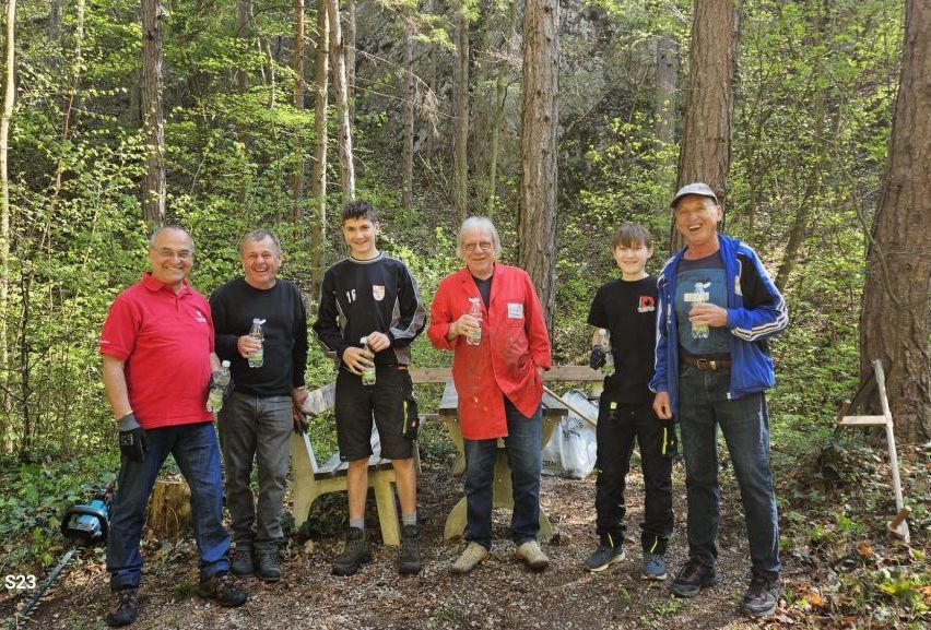Sechs Männer im Wald, lächelnd für ein Foto. Einer hält ein Getränk, andere halten Flaschen. Hinter ihnen steht eine Holzbank und ein Baumstamm.