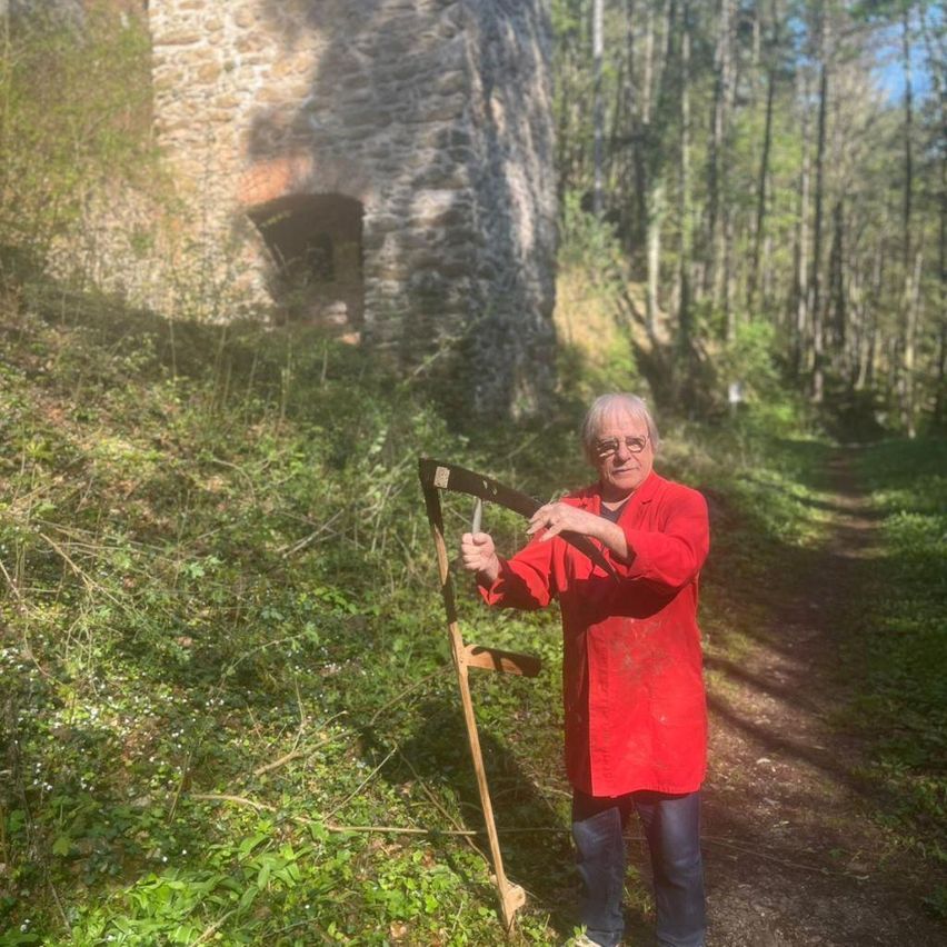 Ein älterer Mann mit Brille und rotem Hemd hält eine Sense im Wald. Dahinter befindet sich ein altes Steingebäude.