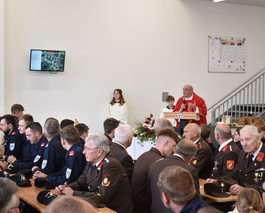 Ein Mann in einer roten Robe steht an einem Rednerpult und spricht eine Gruppe uniformierter Personen an, möglicherweise Feuerwehrleute. Eine Frau steht hinter ihm, und ein Monitor ist an der Wand montiert.