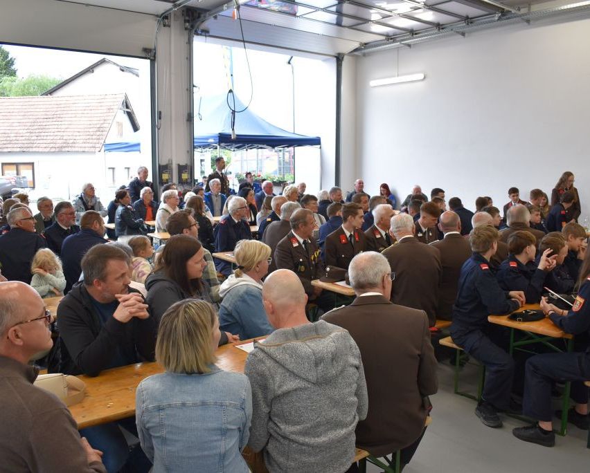 Eine große Gruppe von Menschen sitzt an Tischen in einem Raum mit weißen Wänden und Glasfenstern. Einige tragen Uniformen, während andere in Freizeitkleidung sind. Einige unterhalten sich, während andere auf ihre Telefone schauen.