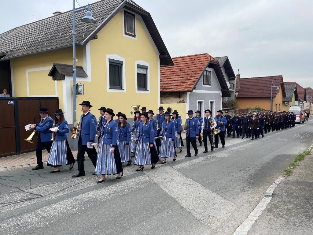 Eine Musikkapelle mit Musikern und einer Gruppe von Menschen in blauen Uniformen läuft eine Straße hinunter, mit Häusern im Hintergrund.