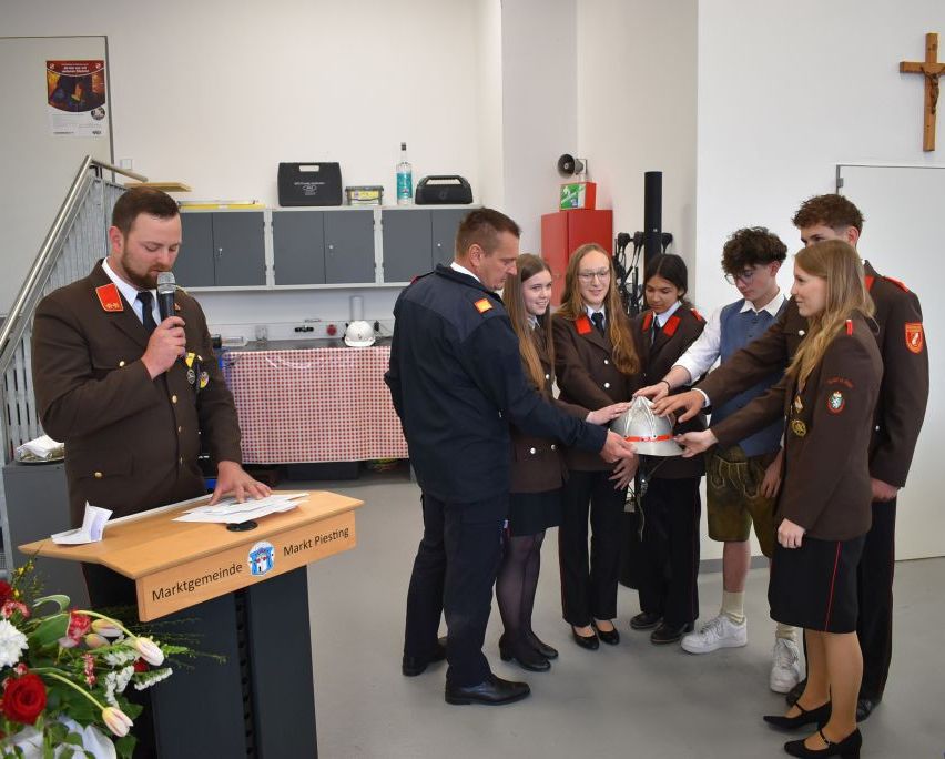 Eine Gruppe von Personen steht in einem Raum, ein Mann mit Mikrofon spricht. Eine Frau in Uniform überreicht einem Mann in Uniform einen Helm.