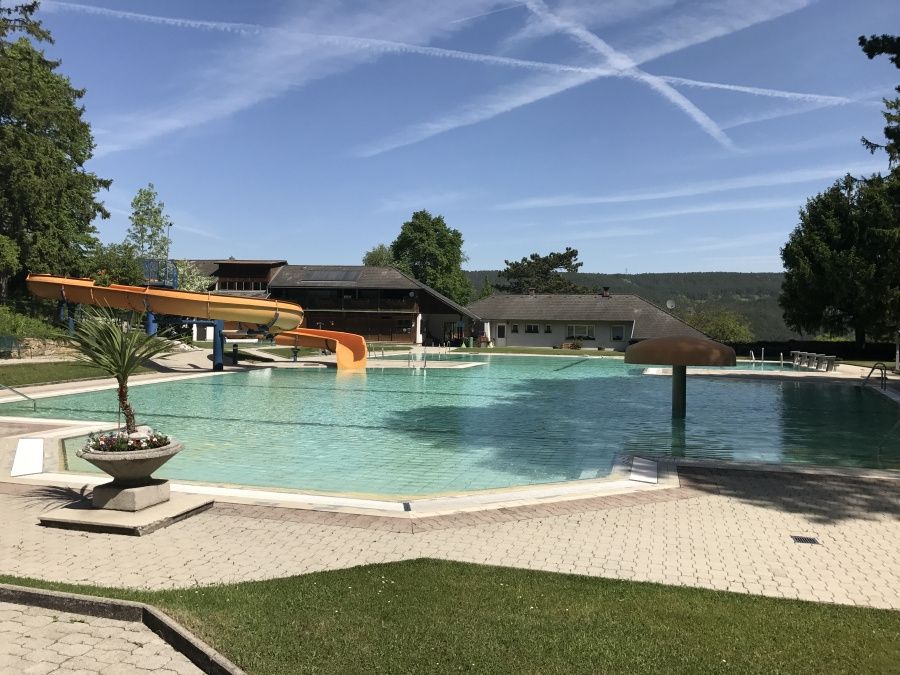 Ein Freibad mit Wasserrutschen und einem klaren blauen Himmel. Bäume und ein Haus sind im Hintergrund.