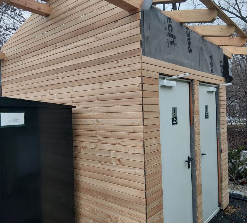 A wooden structure with a gray roof and white doors, surrounded by a wooden fence and a black container.