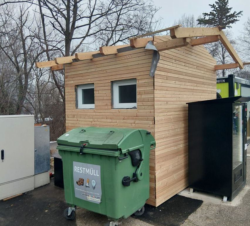 A small wooden house with two windows and a roof is outside. A green trash bin is in front of the house. A black box is beside the house.