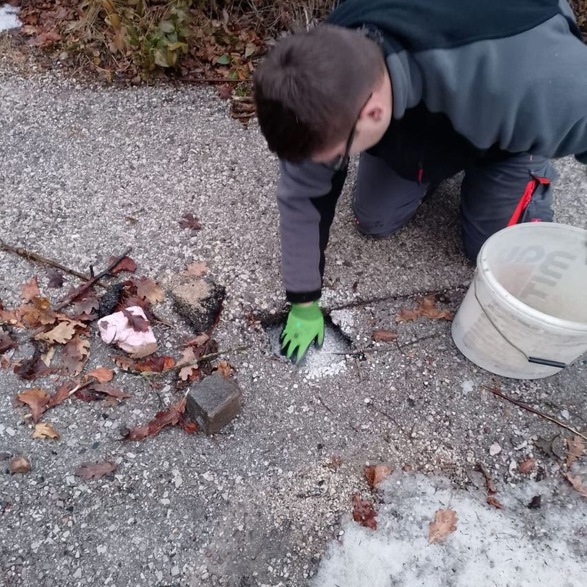 A man wearing gloves is crouched on gravel, spreading salt to melt ice. A bucket is nearby, and leaves are scattered on the ground.