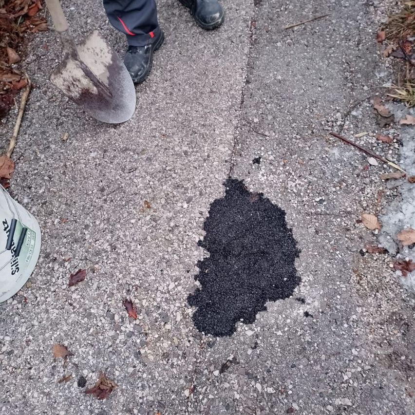 A person wearing dark pants and shoes is digging a hole with a shovel on a concrete road. Nearby, there is a white bucket and a patch of black tar.