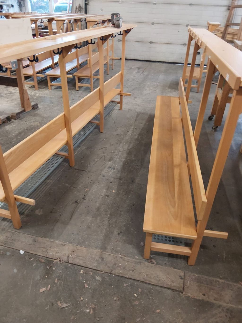 A wooden bench is in a room with a concrete floor. There are several benches in a row. The benches are made of wood and have metal grids on the back.