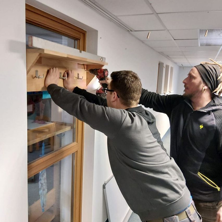 Two men are working together to install a wooden rack with hooks on a wall, one holding the drill and the other guiding the installation.