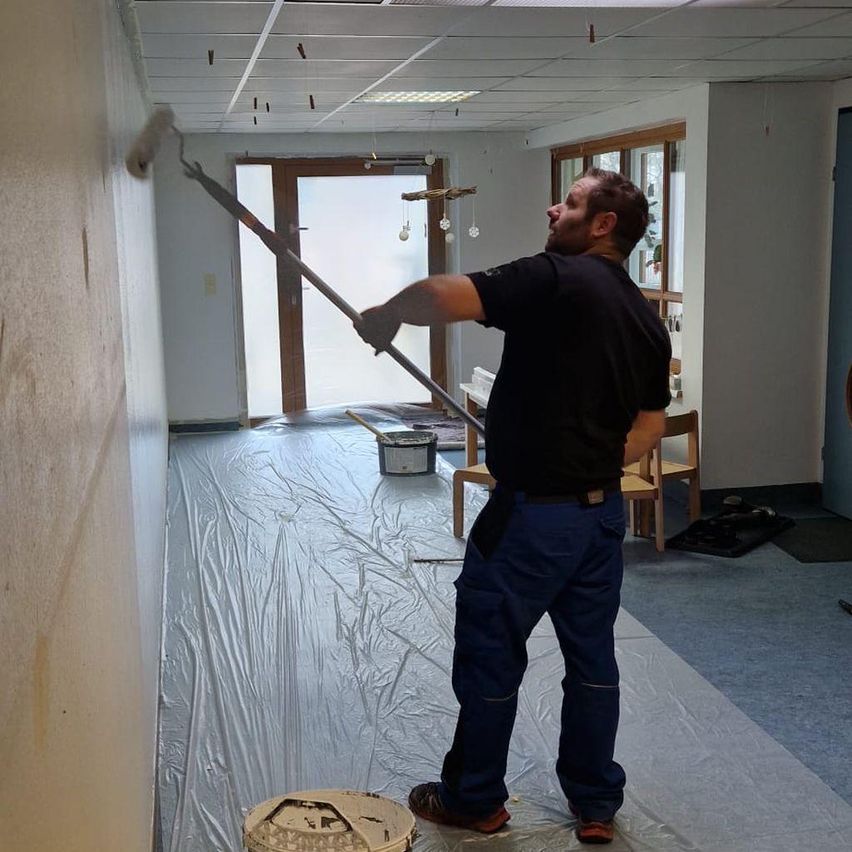 A man in a black shirt and blue pants is painting a wall with a roller. The room has white walls, a wooden door, and a carpeted floor.