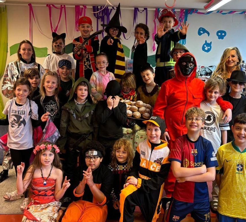 Eine Gruppe von Kindern und Erwachsenen in Kostümen posiert für ein Foto. Einige tragen Superhelden-Umhänge, andere sind als Piraten, Zauberer und Feuerwehrleute verkleidet. In der Mitte steht ein Tisch mit Donuts.