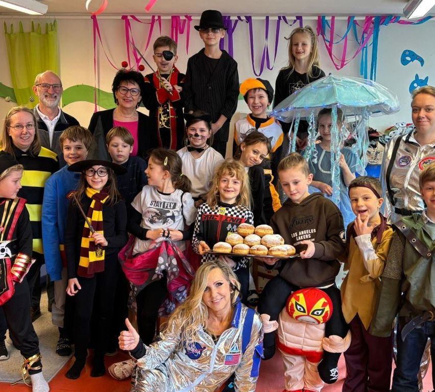 Eine Gruppe von Kindern und Erwachsenen in Kostümen posiert für ein Foto. Einige tragen Gesichtsbemalung, während andere Requisiten halten. Ein Kind hält eine Tablett mit Donuts. Dahinter hockt eine Frau in einer silbernen Astronauten-Anzug und gibt einen Daumen hoch.