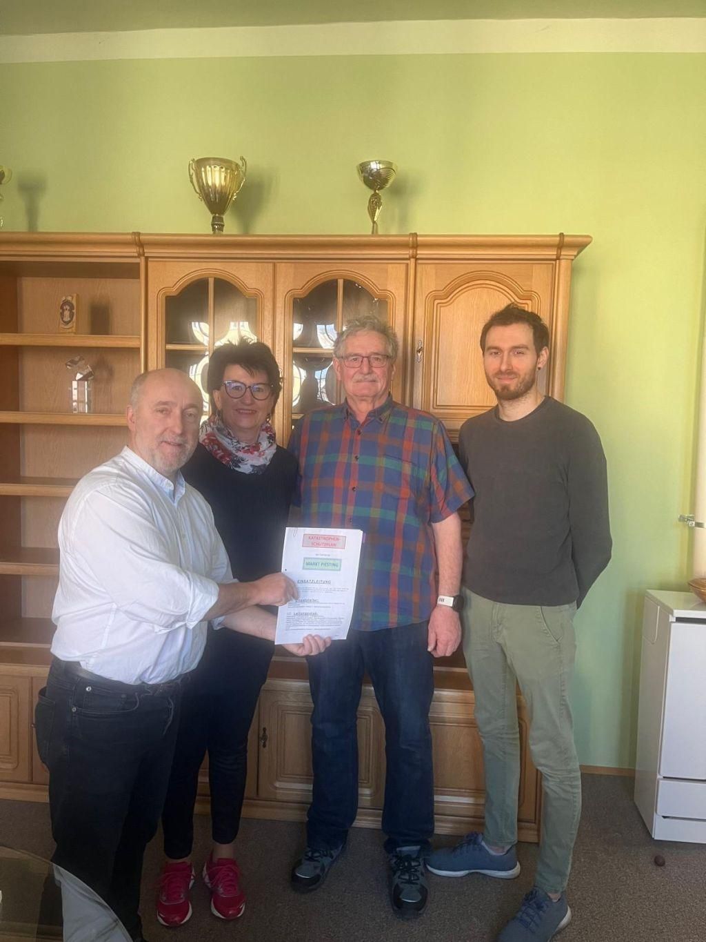 Vier Personen stehen zusammen in einem Raum mit einem hölzernen Schrank dahinter. Sie halten ein Zertifikat. Der Mann auf der linken Seite trägt ein weißes Hemd. Der Mann in der Mitte trägt ein kariertes Hemd. Der Mann auf der rechten Seite trägt ein schwarzes Hemd.