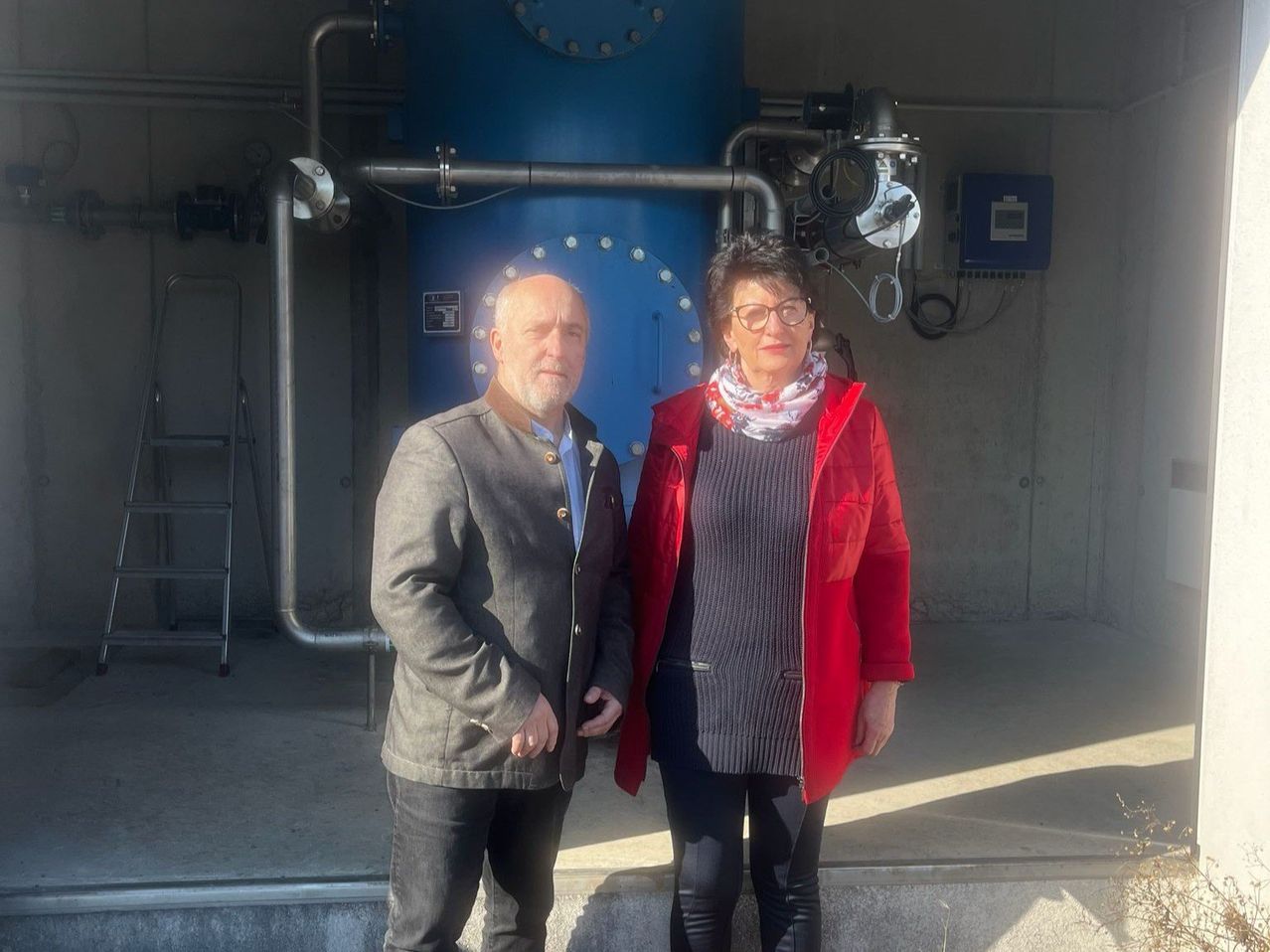 Zwei Personen, ein älterer Mann und eine Frau, stehen vor einem großen blauen Industrietank mit Rohren und Maschinen.