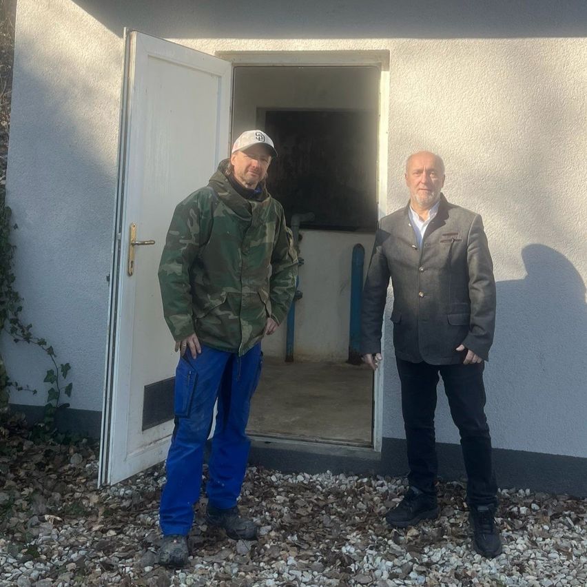 Zwei Männer stehen vor einem Haus mit offener Tür, einer in einer Tarnjacke und der andere in einem grauen Anzug.