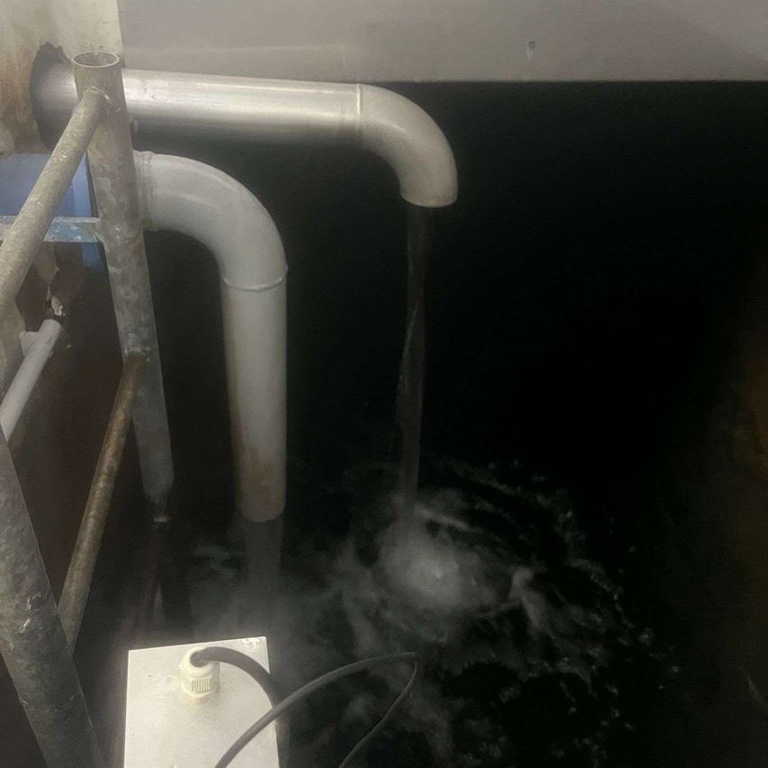 Eine Wasserleitung spritzt Wasser in einen Behälter. Das Wasser spritzt um den Behälter herum. Der Behälter besteht aus Metall.