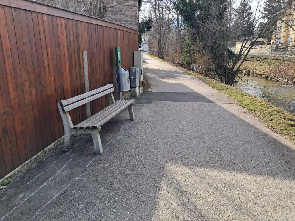 Eine leere Holzbank steht am Rand eines gepflasterten Pfades in der Nähe eines Flusses, mit Mülleimern in der Nähe.