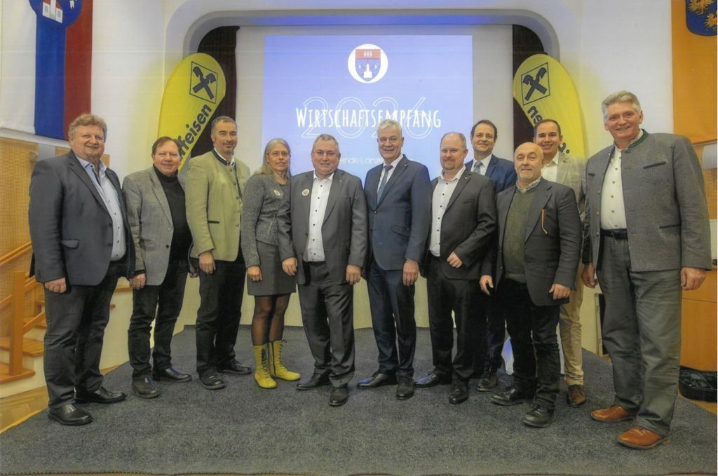 Eine Gruppe von formell gekleideten Personen, die vor einem Bildschirm mit einer Präsentationsfolie stehen. Sie lächeln und posieren für ein Foto.