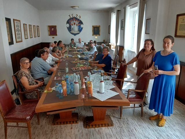 Eine Gruppe von Menschen sitzt um einen langen Tisch in einem Raum, einige sitzen, andere stehen. Der Tisch ist mit verschiedenen Speisen, Getränken und Flaschen beladen. Zwei Frauen stehen am Ende des Tisches, eine hält ein Glas. Der Raum hat einen Holzboden und Wände, die mit gerahmten Bildern geschmückt sind.