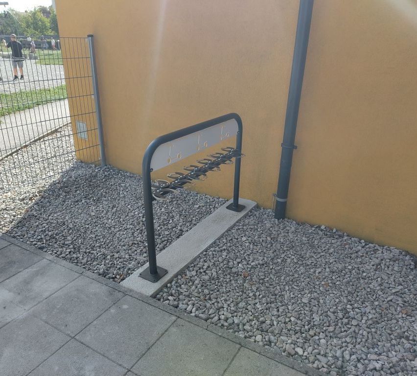 A metal bike rack is mounted on a concrete base and fixed to a yellow wall. Gravel is spread around the base.