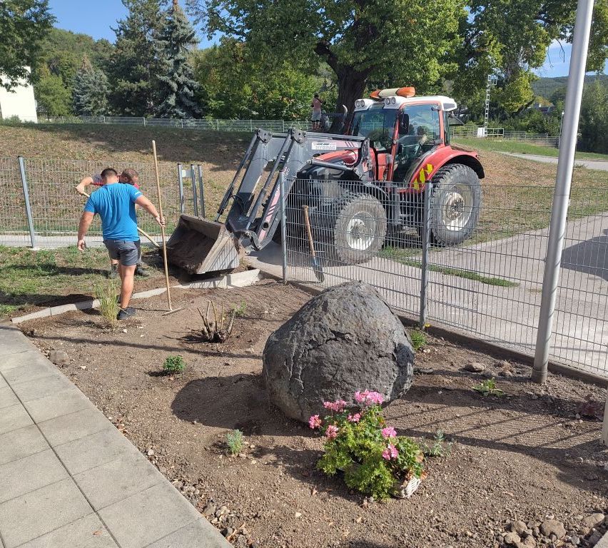 Two men are working on a garden with a large rock, a tractor, and a fence in the background. One man is using a shovel while the other is holding a rake.