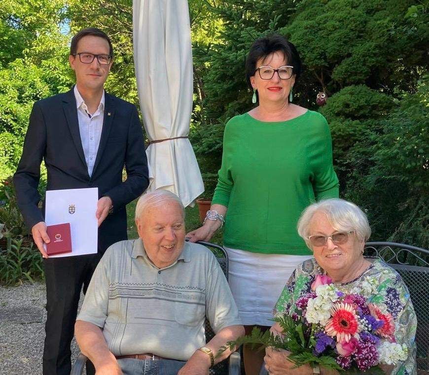 An elderly couple sits on a chair with a man standing behind them. The man holds a red booklet and a piece of paper. The woman holds a bouquet of flowers. The man wears glasses and a suit. The woman wears a green shirt, a white skirt, and earrings. Trees and plants are behind them.