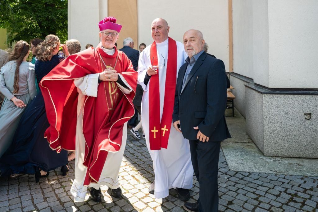 Three men stand outside a church. Two are wearing clerical garb, and one is in a suit. Behind them, people are talking and walking.