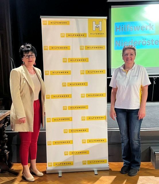 Zwei Frauen stehen neben einem Roll-up-Banner mit dem Text 'Hilfswerk Niederösterr.' darauf und lächeln in die Kamera.