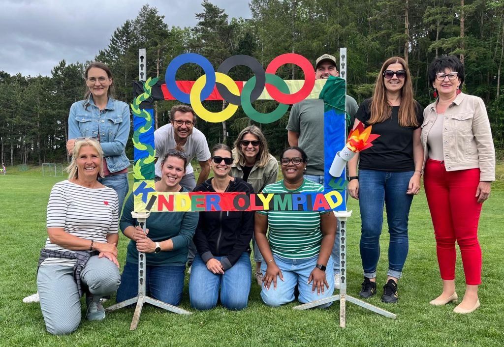 Eine Gruppe von Menschen posiert für ein Foto vor einer selbstgemachten olympischen Kulisse. Sie lächeln und einige tragen Sonnenbrillen. Dahinter stehen Bäume und ein bewölkter Himmel.