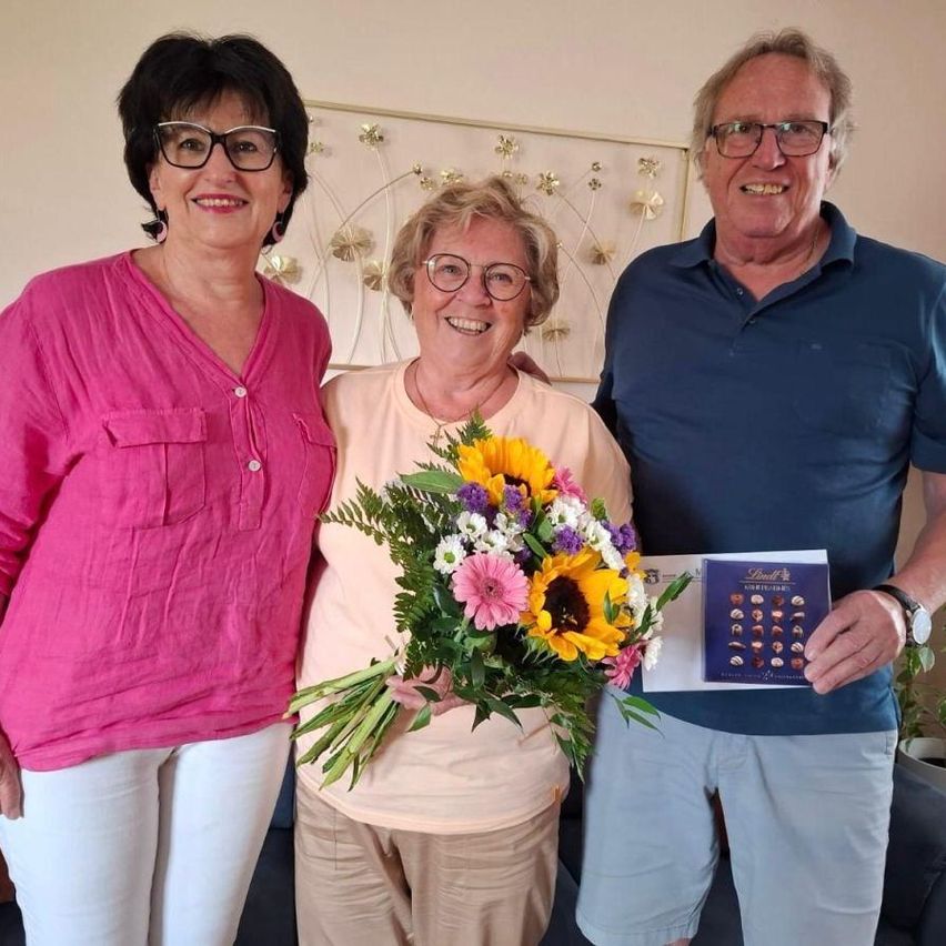 Three smiling adults pose for a photo with a bouquet of flowers. The woman in the middle holds the bouquet, while the man on the right holds a card.