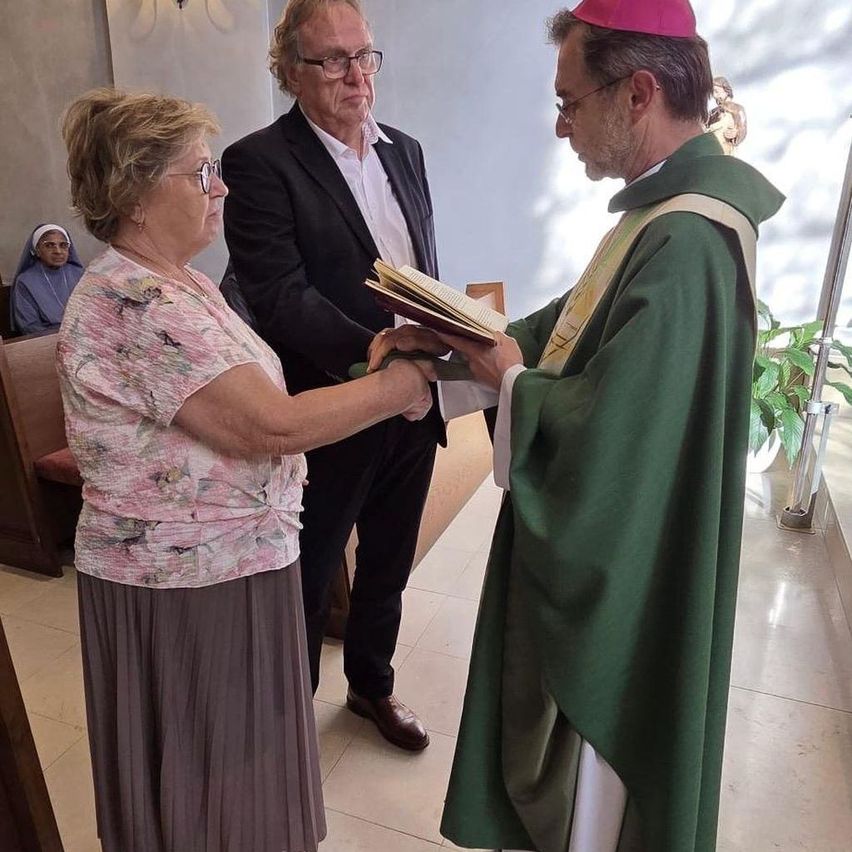 A priest hands a book to an older woman in a floral blouse. A man in glasses stands behind them. They are in a chapel with white walls and a nun in the background.
