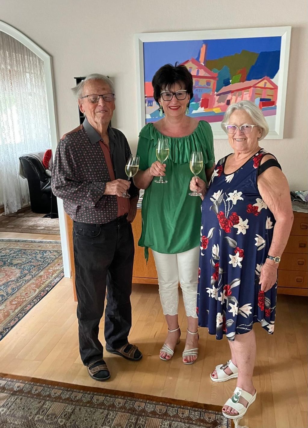 Three people stand together in a living room, smiling for a photo. Two women hold wine glasses, and one man has a glass. Behind them is a painting on the wall and a rug on the floor.