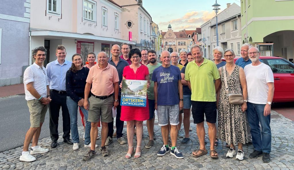 A group of people stands on a cobblestone street, smiling. A woman holds a sign that reads 'Ortskern-Entwicklung'. Buildings, a tower, and street lights are in the background.