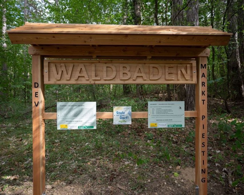 A wooden signboard reads 'WALDBADEN'. Two informational signs hang on the sides, one mentioning 'Stoltz' and the other 'Dorf!'. The signboard stands in a forest area.