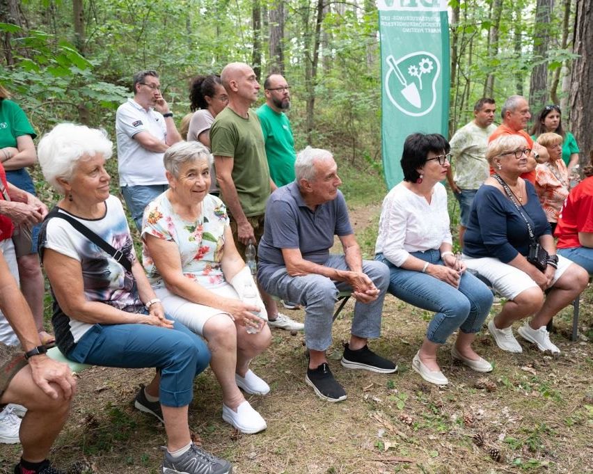 A group of older people are sitting on a bench in a forest. Some are looking at the camera. A green banner is placed behind them.