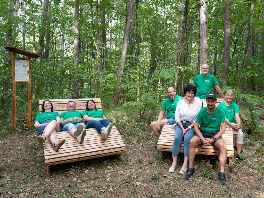 A group of people in green shirts are sitting on wooden benches in a forest. They are all smiling and posing for a picture.