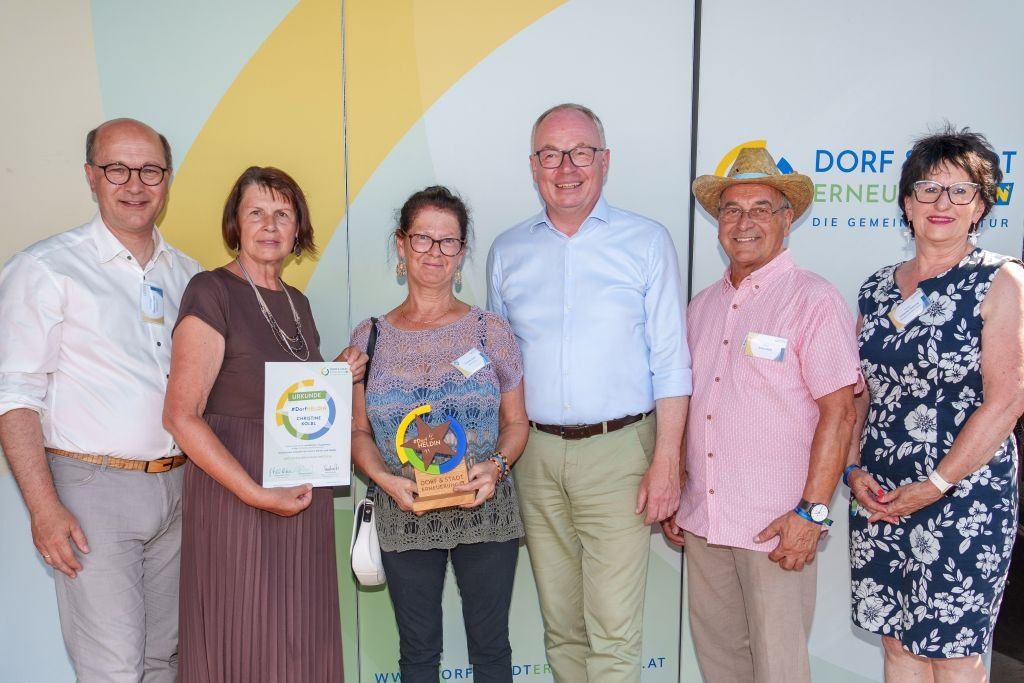 Five people pose for a photo. One woman holds an award and a certificate. Another woman holds a trophy. A man in a straw hat stands on the right. They stand in front of a wall with the word Dorferneuert.