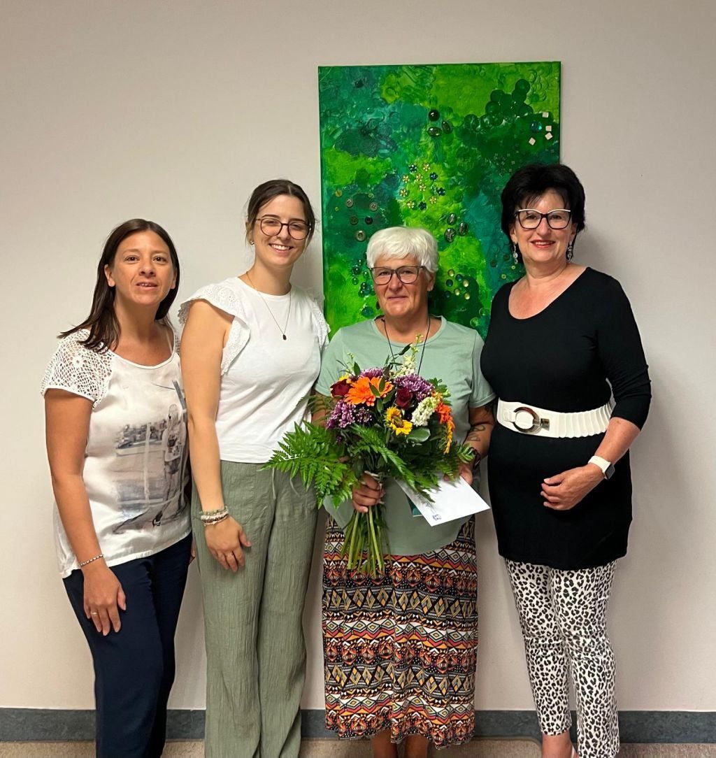 Four women stand together in front of a green painting, smiling for a photo. The woman in the center holds a bouquet of flowers.