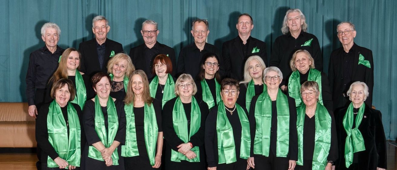 Ein Chor von Erwachsenen in schwarzer Kleidung und grünen Schals posiert für ein Foto auf der Bühne vor einem grünen Vorhang.