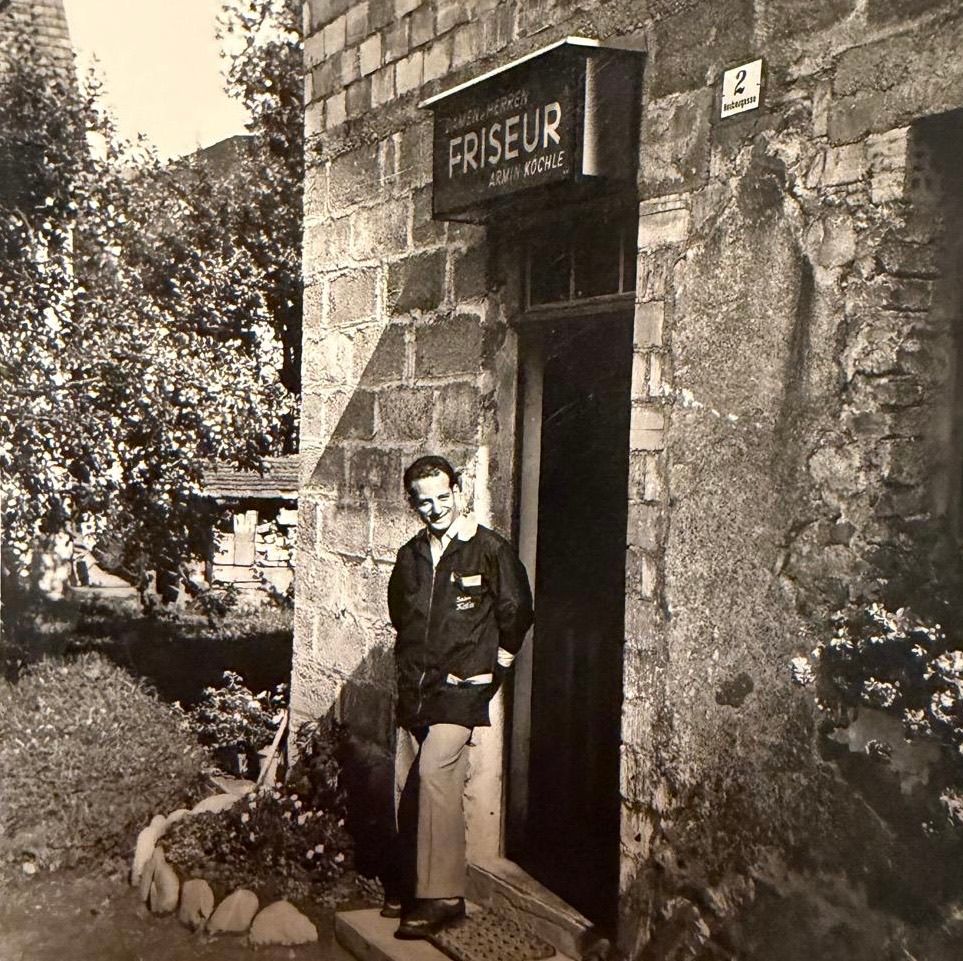 Ein altes Schwarz-Weiß-Foto zeigt einen Mann vor der Tür eines Steingebäudes mit einem Schild, das Friseur besagt.