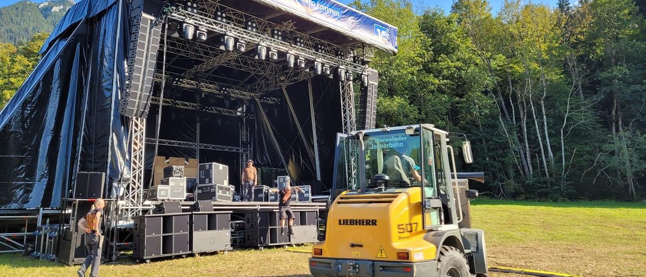 A setup for an outdoor event with a stage featuring a banner reading Morgenland Bochum. Two individuals are working on the stage, one is jumping and the other is standing. A yellow Liebherr forklift is positioned in front of the stage.