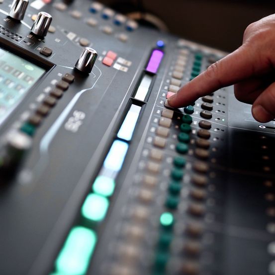 A hand is pressing a red button on a sound mixing console. The console has a digital display and various colored lights and knobs.