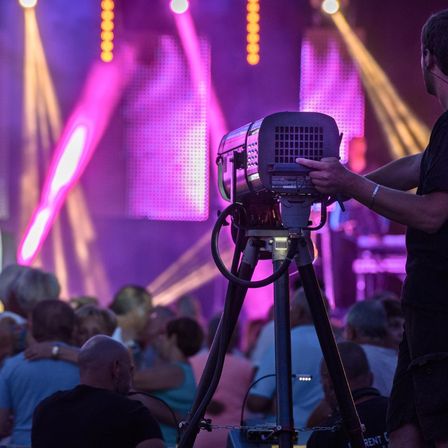 Ein Konzert ist im Gange, wobei eine Person eine Kamera auf einem Stativ hält und das Publikum vor einer Bühne mit leuchtendem Licht einfängt.