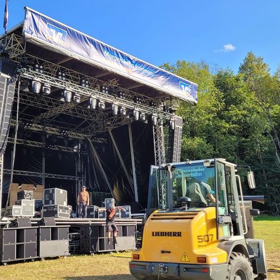 A stage setup with speakers and equipment is prepared for an event. Two individuals are on the stage, with one performing a backflip. A yellow Liebherr vehicle is parked beside the stage.