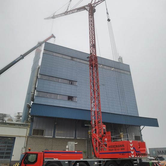 Ein Liebherr Mobilkran mit ausgefahrenem Ausleger steht vor einem großen Industriegebäude mit blauer Fassade. Ein weiterer Kran ist dahinter positioniert.